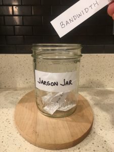 Image of hand depositing hand-written note that says 'bandwidth' into the Jargon Jar